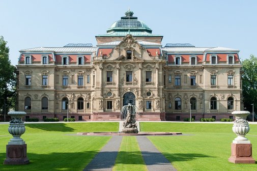 Bundesgerichtshof Blick auf einen im Stil des Neobarocks gestaltetes Palastgebäude im Sonnenschein mit großen Rasenflächen und einem Brunnen davor.