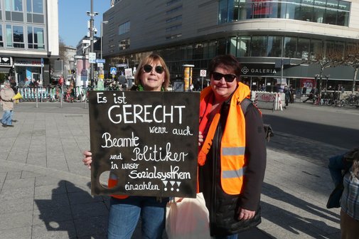 Aktion zum Equal Pay Day 2026 in Frankfurt An der Hauptwache in Frankfurt halten bei strahlendem Sonnenschein zwei Frauen mit Sonnenbrillen ein Schild mit der Beschriftung "Es ist gerecht, wenn auch Beamte und Politiker in unser Sozialsystem einzahlen!" hoch.