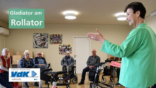 Eine Frau in hellgrünem Pulli steht vor einer Gruppe Senioren und Seniorinnen und erklärt die Funktionalität eines Rollators.