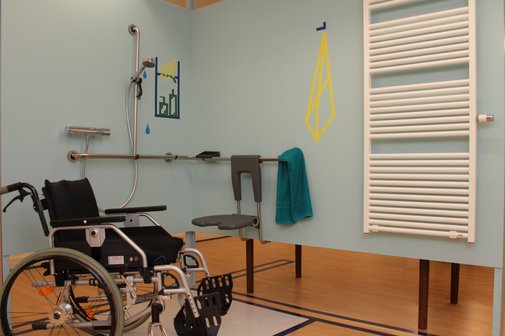 Bad in der Ausstellung Hallo Freiheit! Barrierefreies Badezimmer mit Duschbereich, Haltegriffen und Duschsitz; daneben steht ein Rollstuhl, an der Wand hängt ein Handtuch und ein Heizkörper ist zu sehen