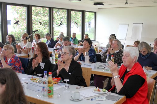 Seminarraum mit langen Tischreihen und großen Fenstern an der einen langen Seiten, gut gefüllt, mit vielen Frauen, die interessiert zuhören.