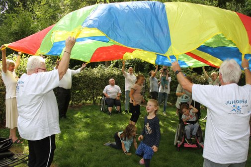 Erwachsene und Kinder heben in einem Park ein großes, buntes Schwungtuch, während Kinder darunter spielen.